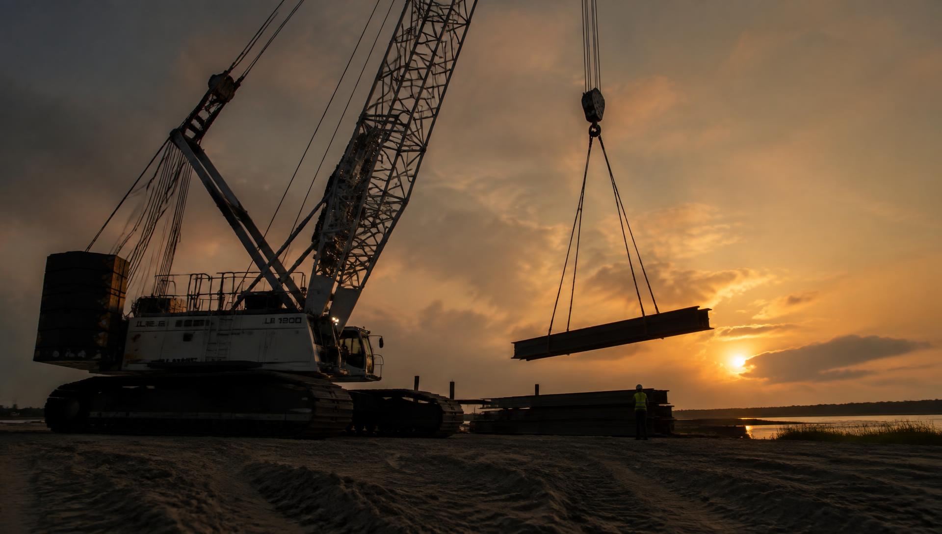 Crane lifting steel at a coastal construction site at sunset
